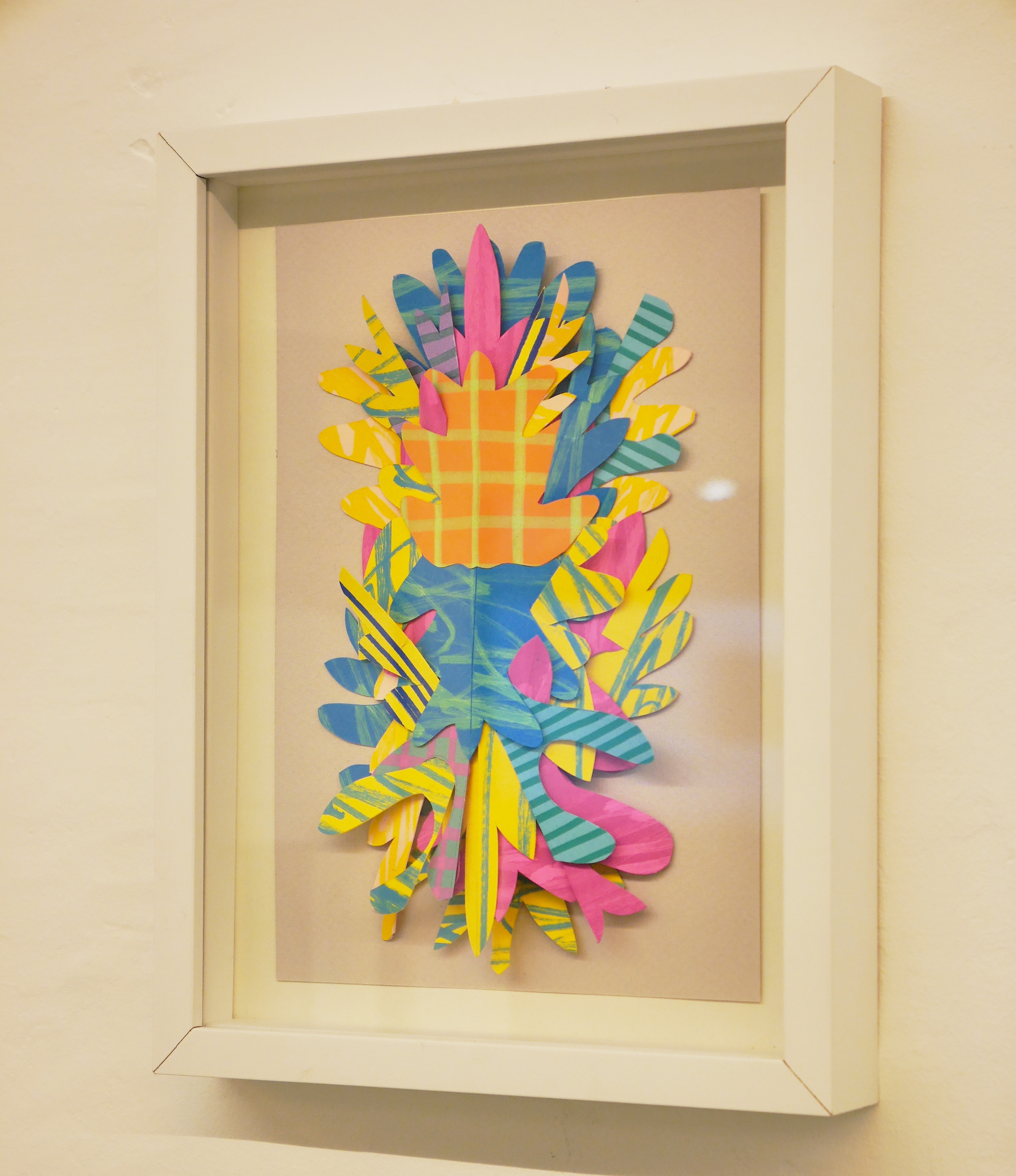 Paper artwork, framed and hanging on the wall. The artwork resembles an organic floral form, with similar silhouettes layered on top of each other in vibrant colors.